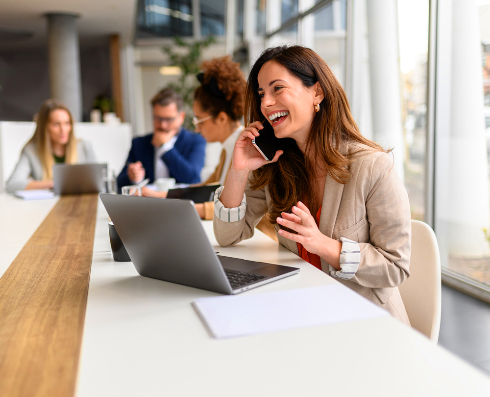 A woman smiling while talking on phone, sitting at a desk with a laptop in an office setting.