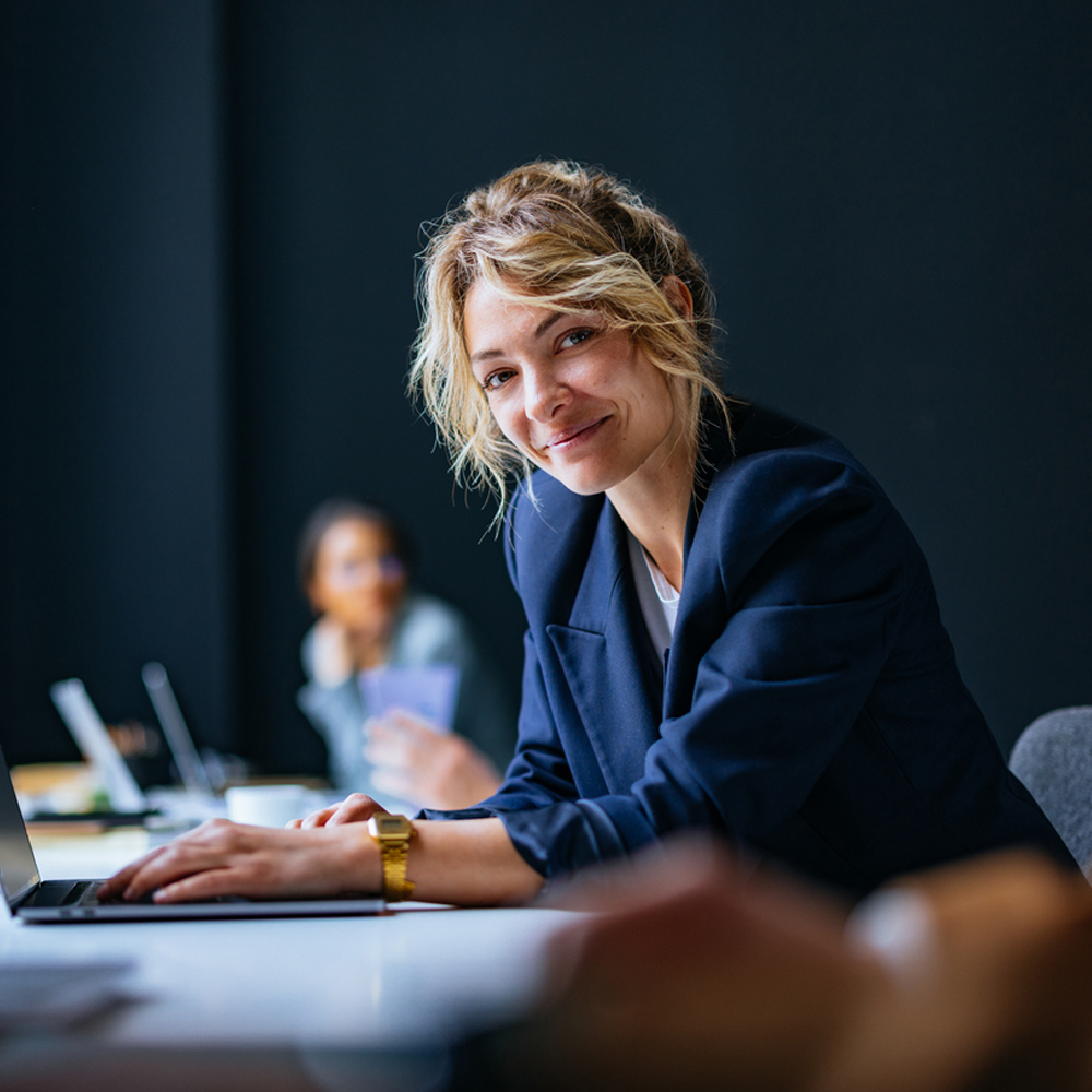 A woman in a blue blazer smiling while working on a laptop in an office setting.