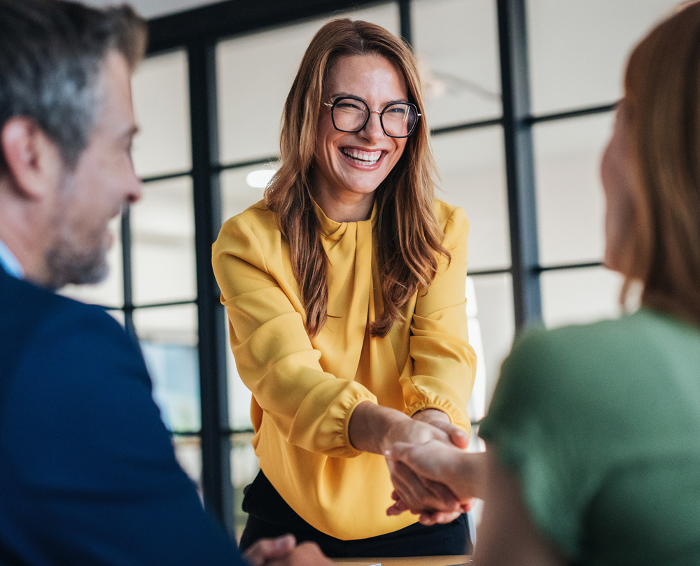 A woman in a yellow blouse smiles while shaking hands in an office setting.