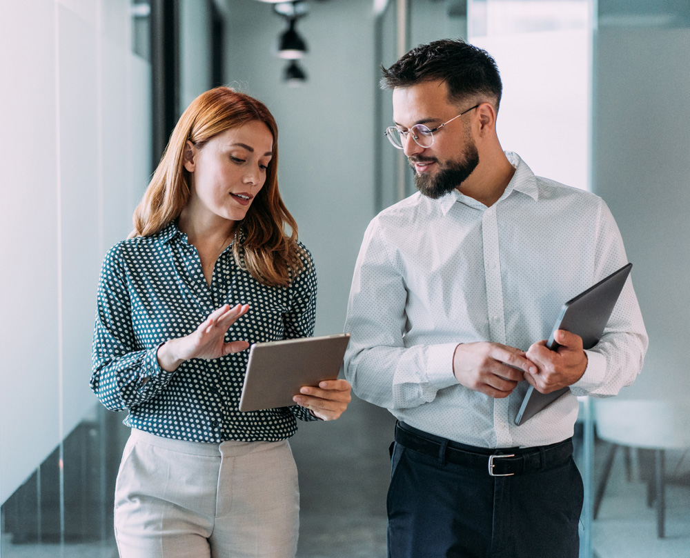 A woman and a man discussing work while holding tablets in an office hallway.