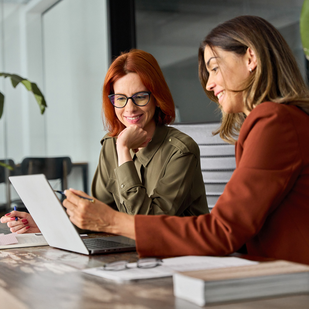 Two women smiling while collaborating on a laptop in an office setting.