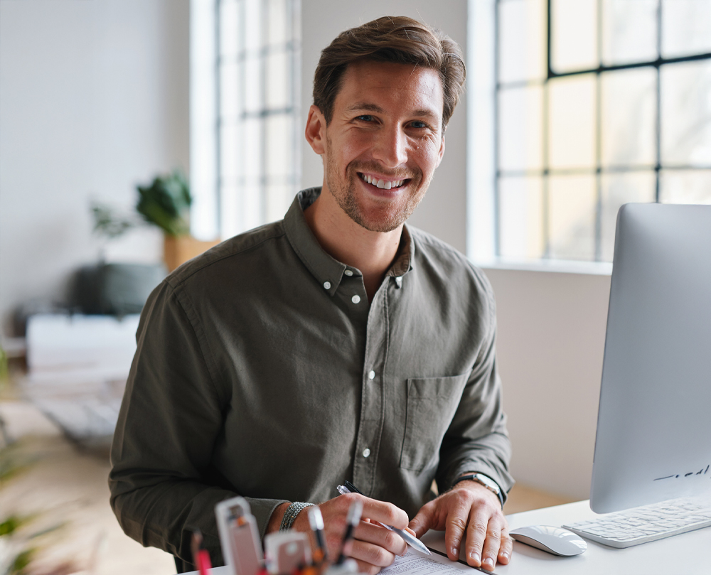 A man smiling and writing at a desk with a computer in a bright office.