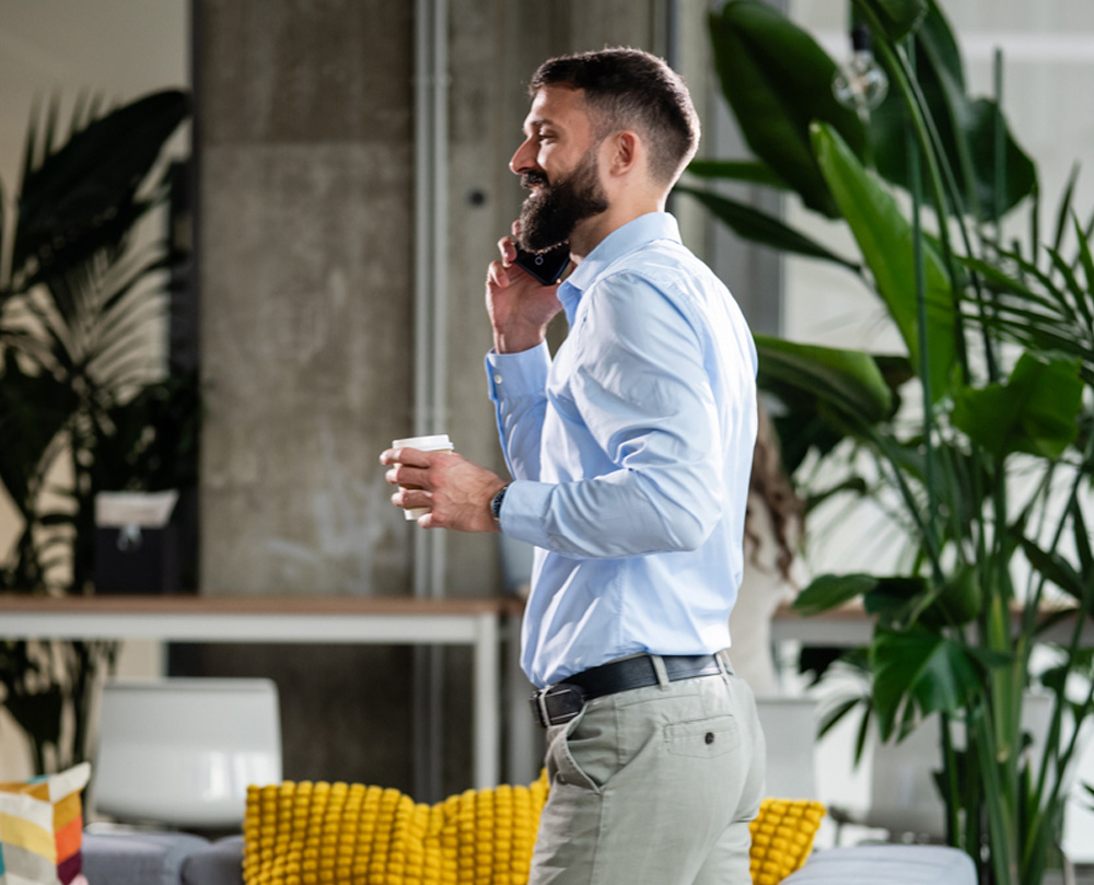 A person in a blue shirt talking on the phone while holding a coffee cup, surrounded by indoor plants.