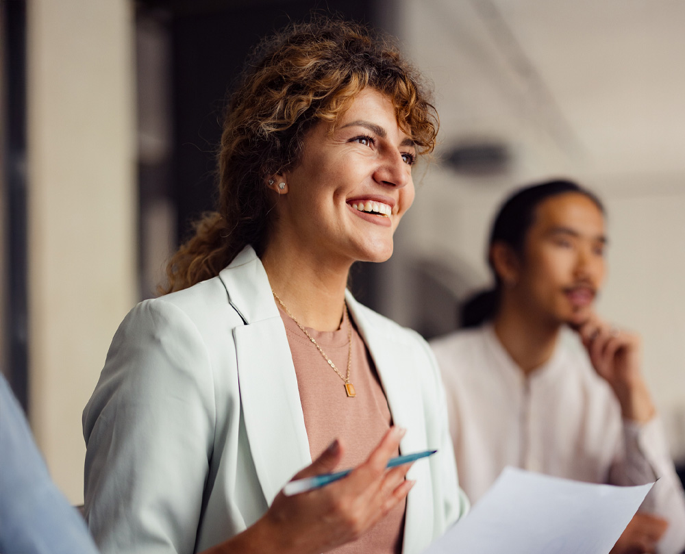 A woman smiling and holding papers during a meeting, with a man in the background.